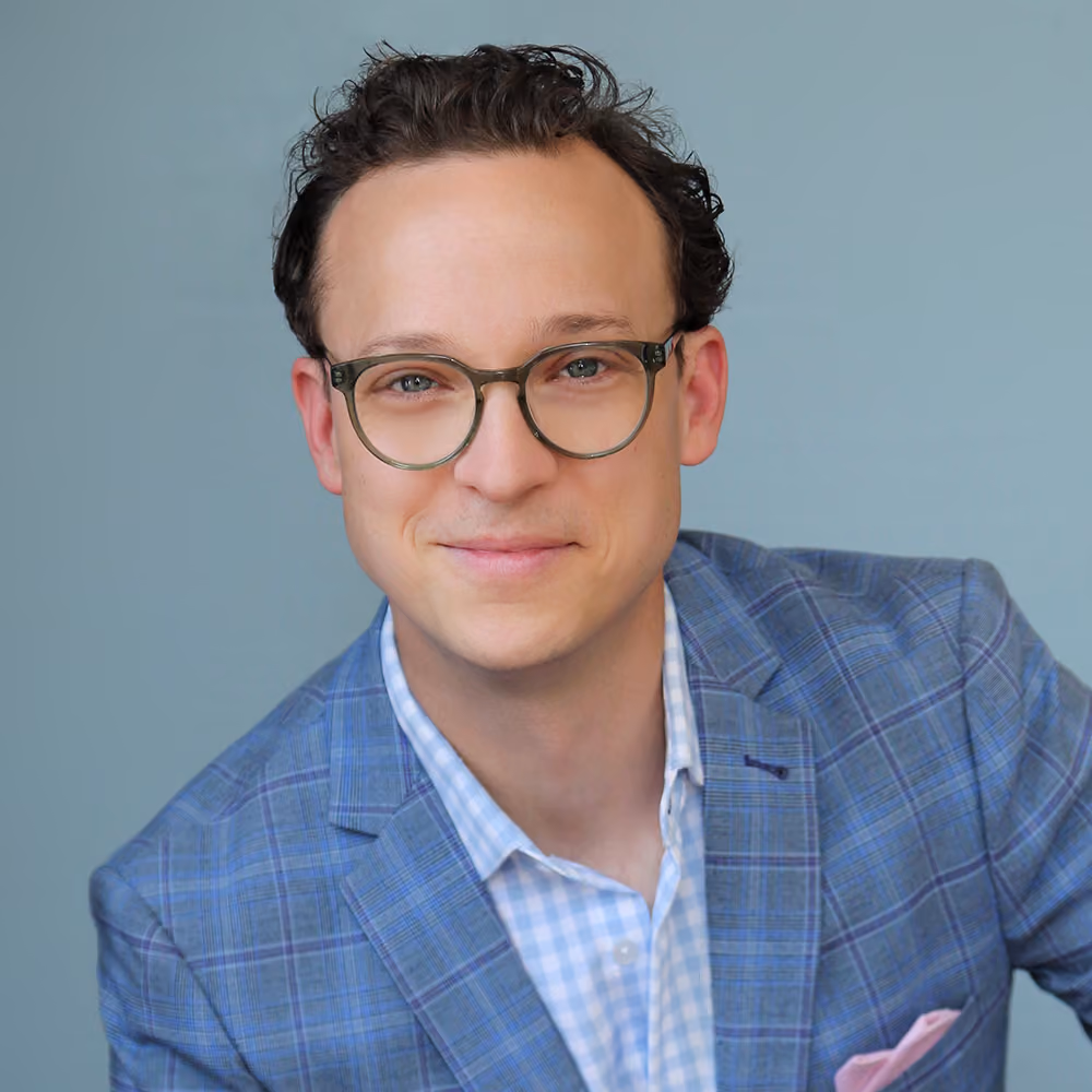 Smiling man with glasses wearing a blue checkered blazer and light blue checkered shirt, against gray background.