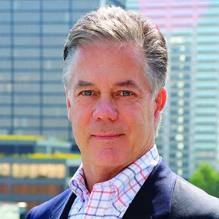 Middle-aged man with gray hair wearing a checked shirt and dark blazer in an urban outdoor setting.