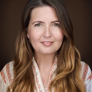 Portrait of a woman with long brown hair wearing a patterned blouse on a dark background.