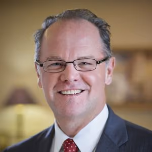 Smiling middle-aged man wearing glasses, a suit, white shirt, and red tie in an indoor setting.