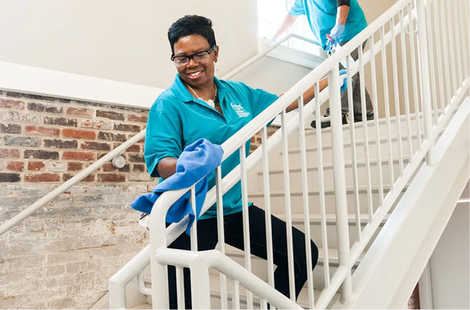 Two people in teal shirts wearing gloves cleaning white stair railings in a brick-walled building.