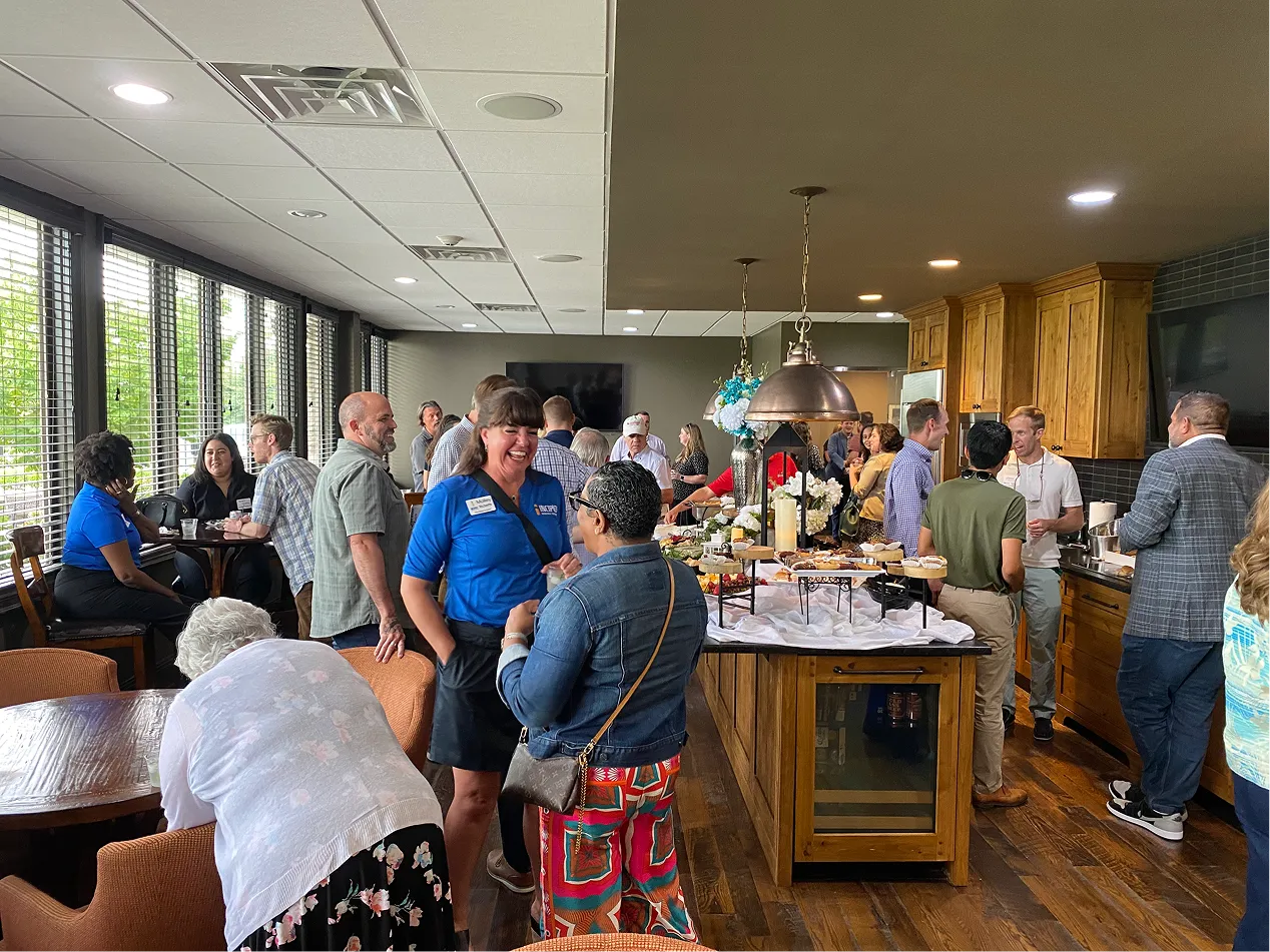 People socializing around a buffet table in a well-lit room with large windows and wooden cabinets.