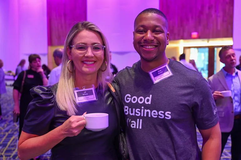 Smiling woman holding a coffee cup and man wearing a Good Business Y’all shirt at a social event.