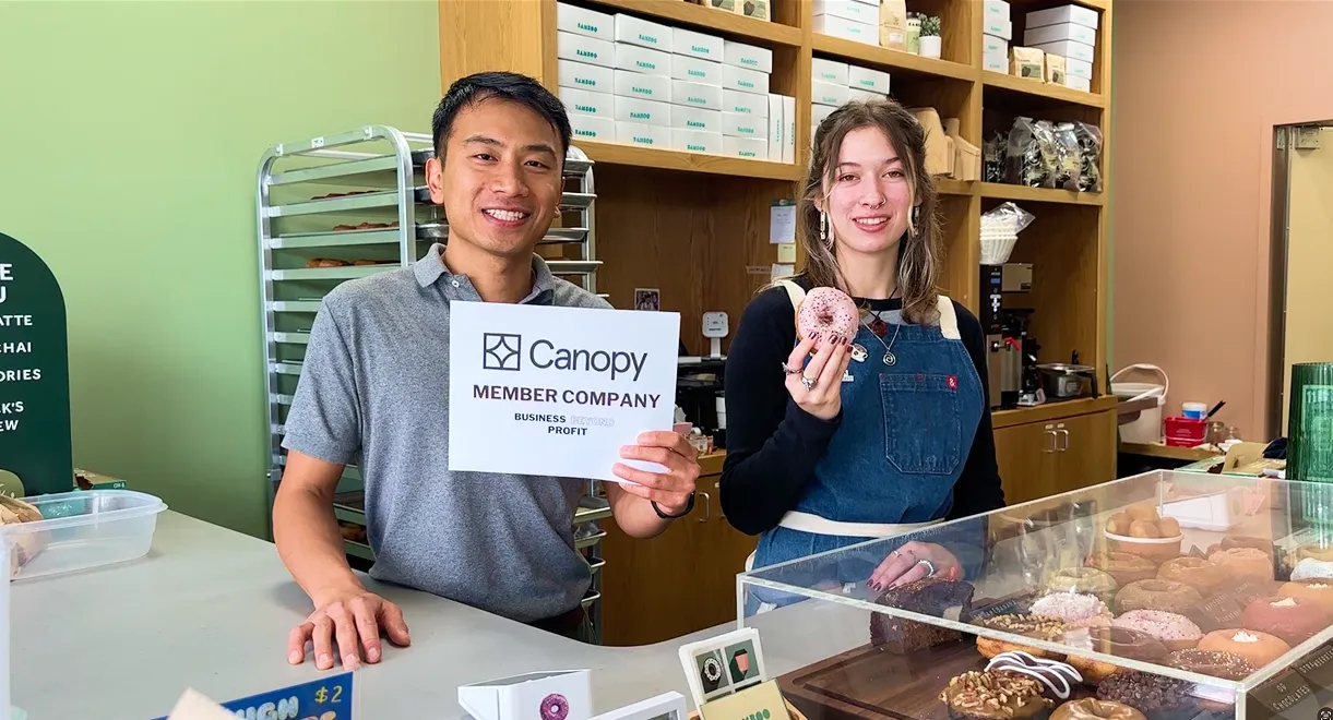 Two bakery workers behind counter; man holds Canopy member sign, woman holds a pink sprinkled donut.