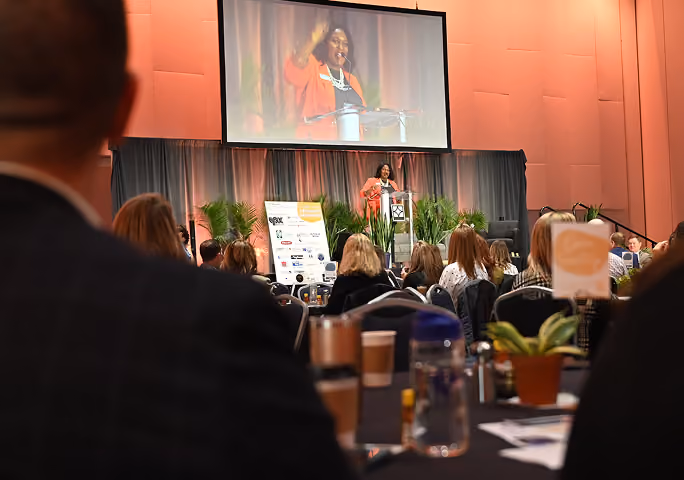 Woman speaking at podium on stage in front of seated audience with her image on large screen