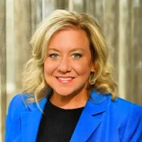 Smiling woman with blonde hair wearing a blue blazer and black top in front of a blurred wood background.