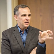 Man in a suit jacket gesturing with his right hand while speaking indoors.