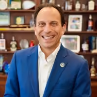 Smiling man in blue suit and white shirt standing in front of a decorated wooden shelf.