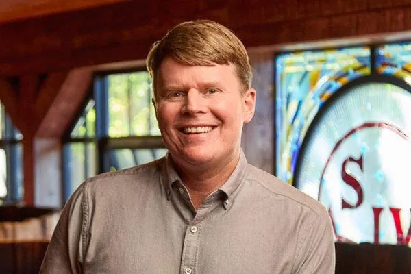 Smiling man with light brown hair wearing a gray button-up shirt in a warmly lit room.