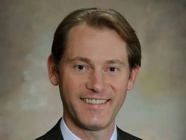 Smiling man with light brown hair wearing a dark suit and light blue shirt.