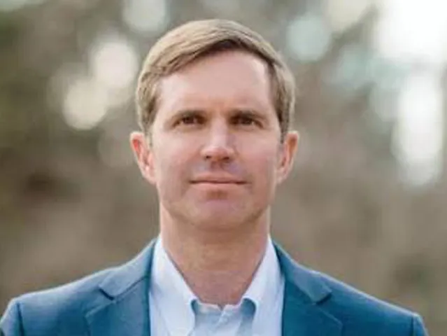 Man with short light brown hair wearing a blue suit jacket and light blue shirt outdoors.
