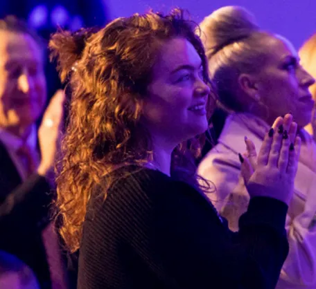 Smiling woman with curly hair clapping among a crowd at an event with purple lighting.
