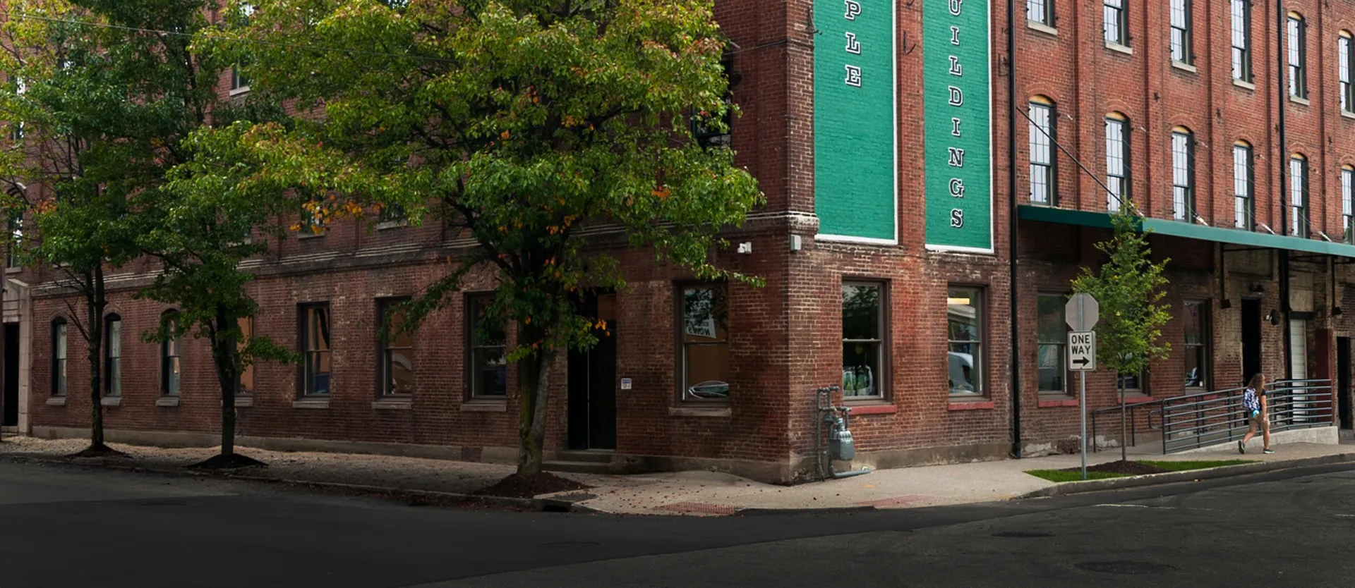 Brick building with green signs, trees on sidewalk, and a person walking near a one way street sign.