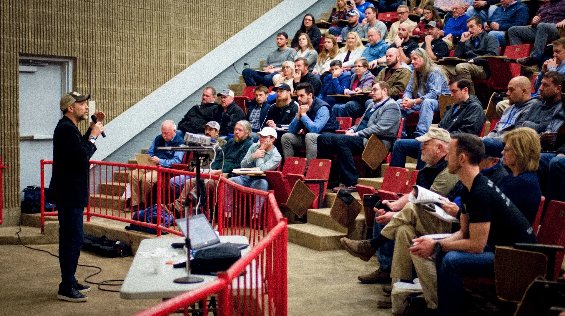 Man with microphone speaking to a seated audience in a lecture hall with red chairs and railings.