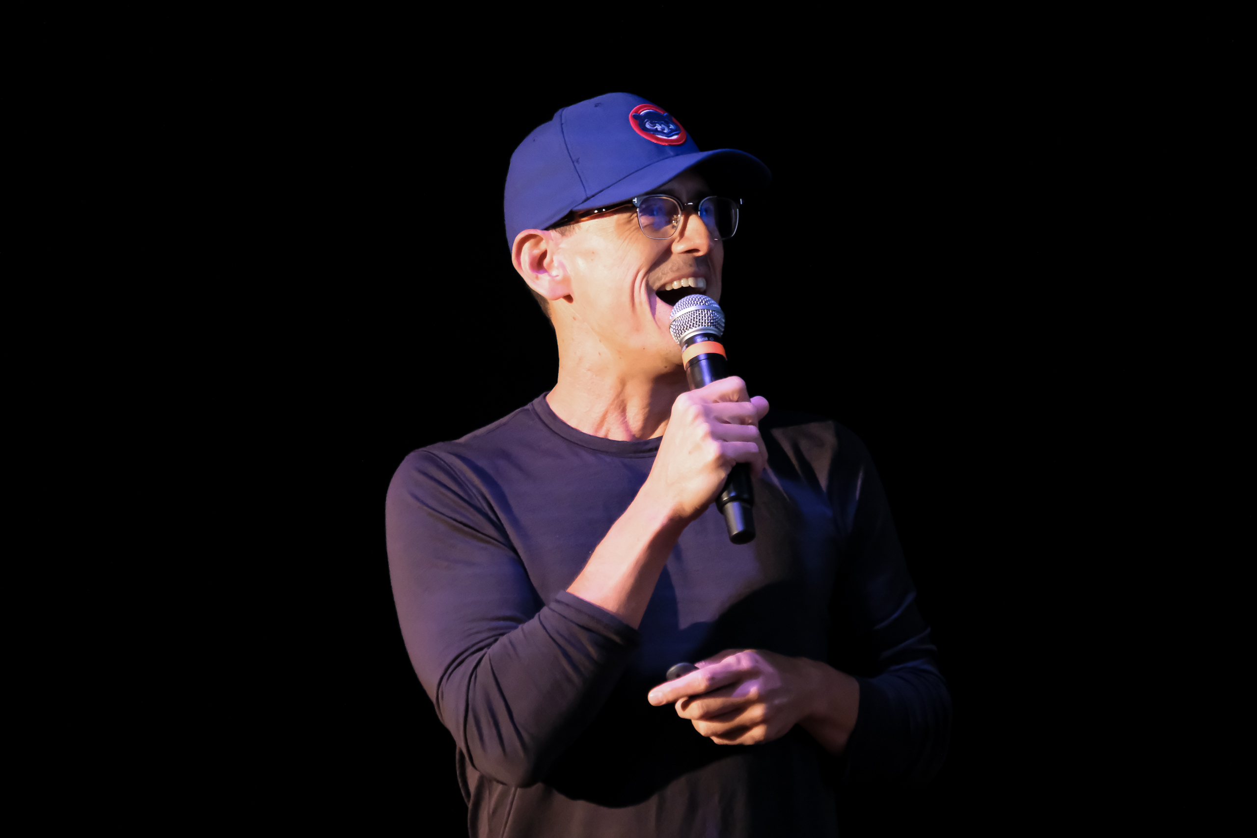 Man wearing glasses and a blue Chicago Cubs cap speaking into a microphone against a black background.