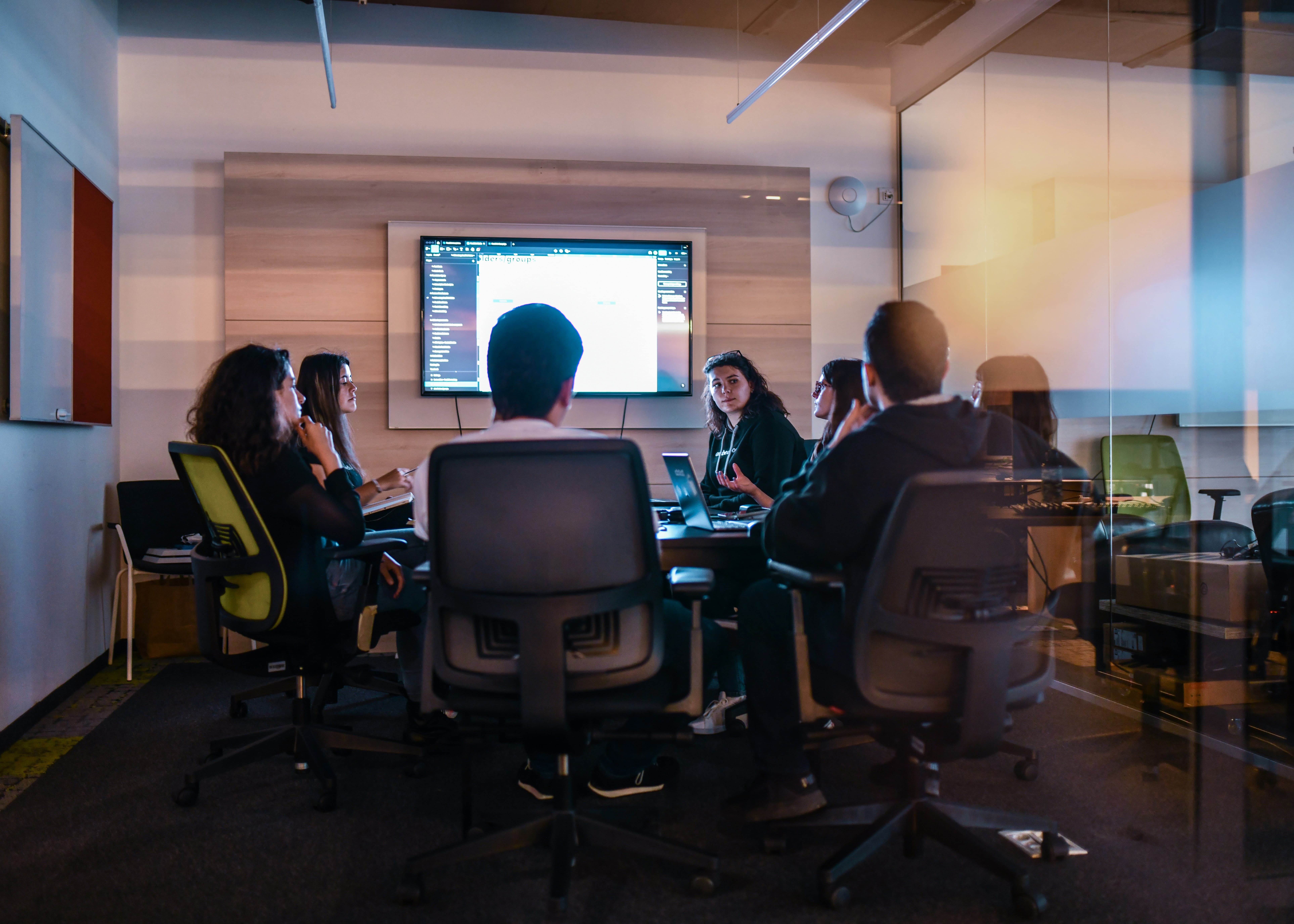 Group of people sitting around conference table watching a presentation on a wall-mounted screen.