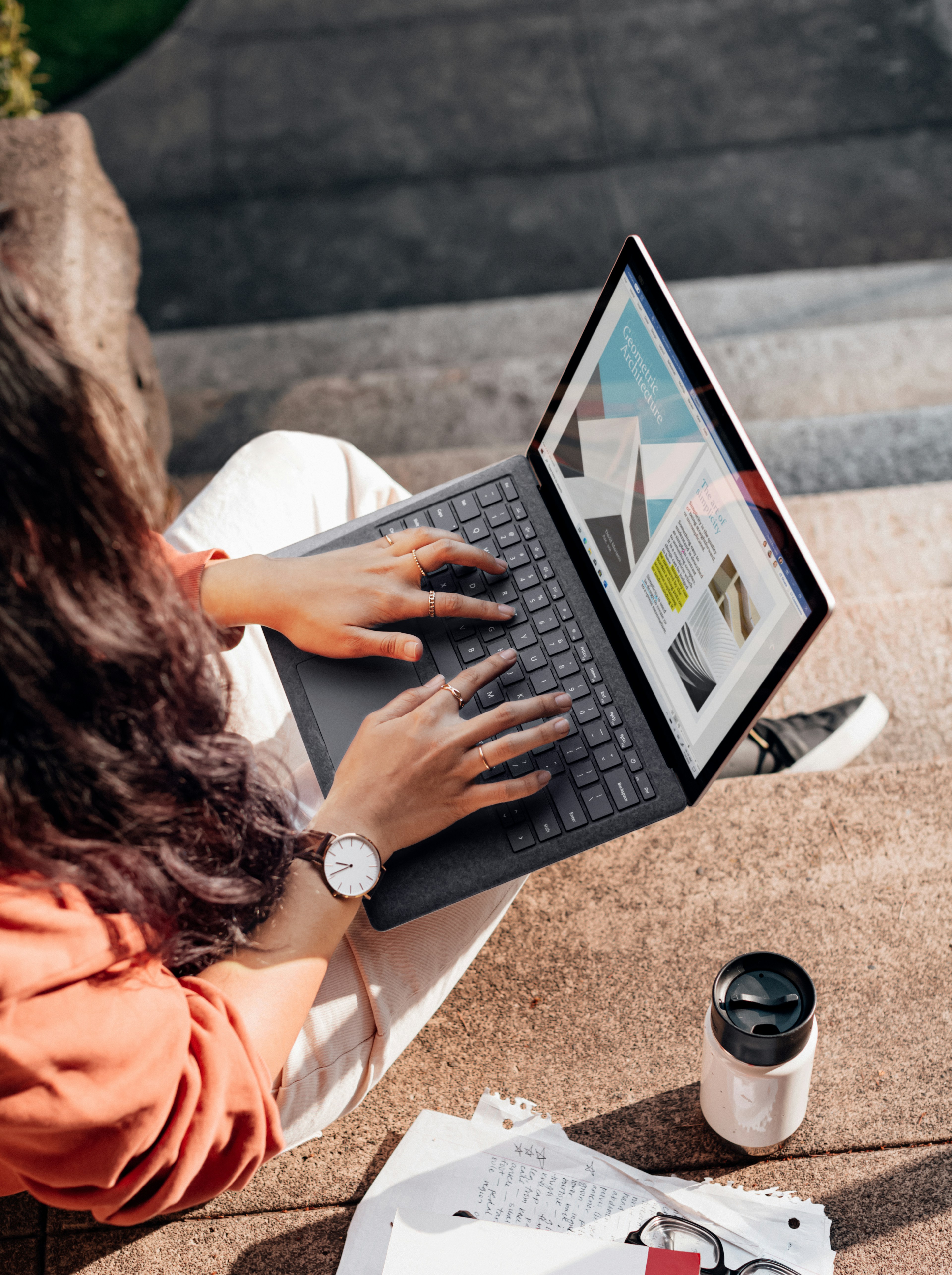 Person sitting on outdoor steps typing on a laptop showing a geometric architecture webpage.