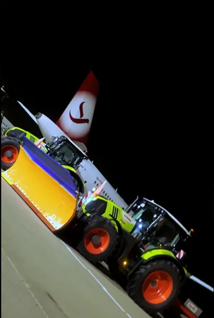Zwei mit Schneepflügen ausgestattete Traktoren stehen nachts auf dem Flughafen vor einem Flugzeug mit rotem Logo am Heck.