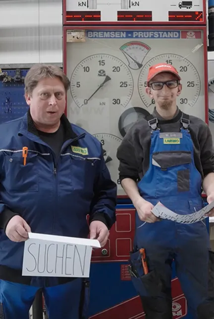Zwei Männer in Arbeitskleidung stehen vor einem Bremsprüfstand, einer hält ein Schild mit der Aufschrift 'SUCHEN'.