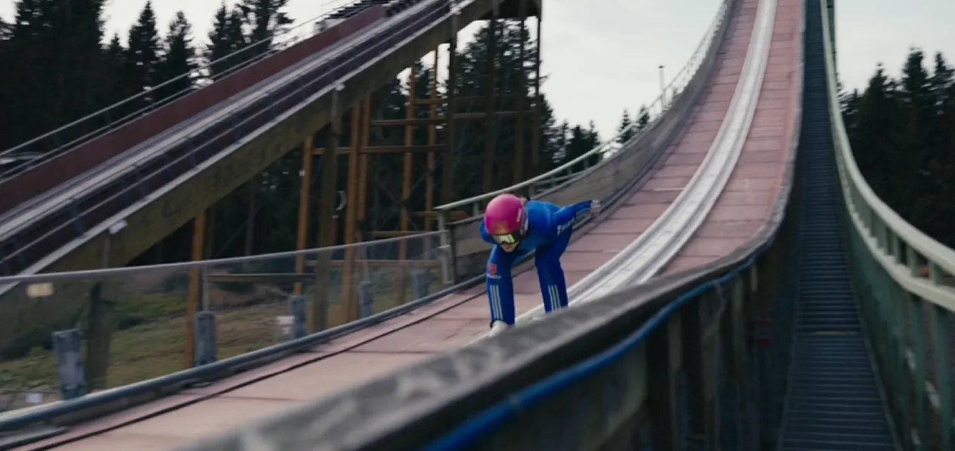 Skispringer in blauer Ausrüstung und rosa Helm fährt eine Schanze hinunter.