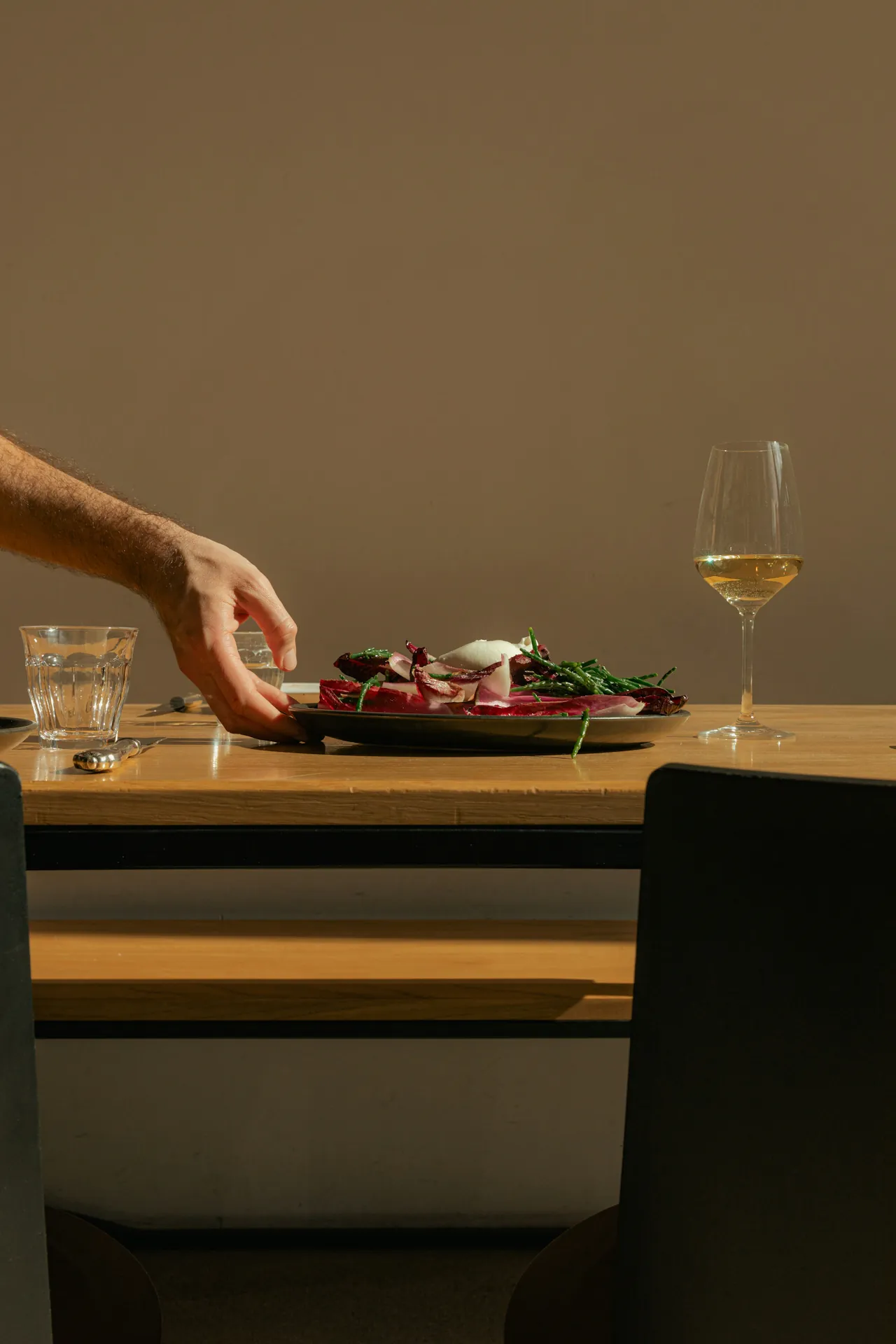 A man's hand reaches across a warm, candlelit wooden dining table to adjust a dark plate of dressed radicchio salad with green beans and burrata, alongside a water glass and a stemmed glass of white wine.