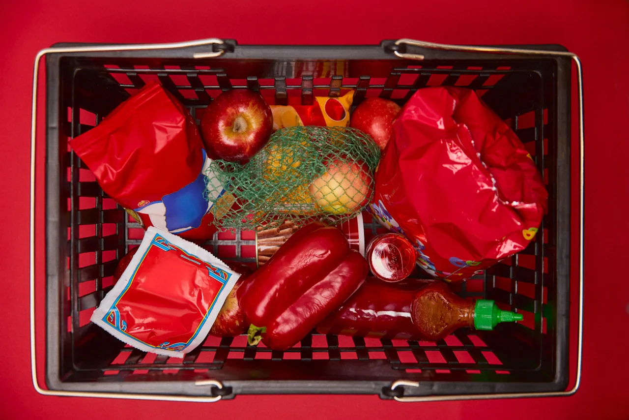 A top-down view of a black plastic shopping basket filled with red apples, red bell peppers, a green mesh bag of citrus, red packaged snacks, and a condiment bottle, set against a vivid red background.