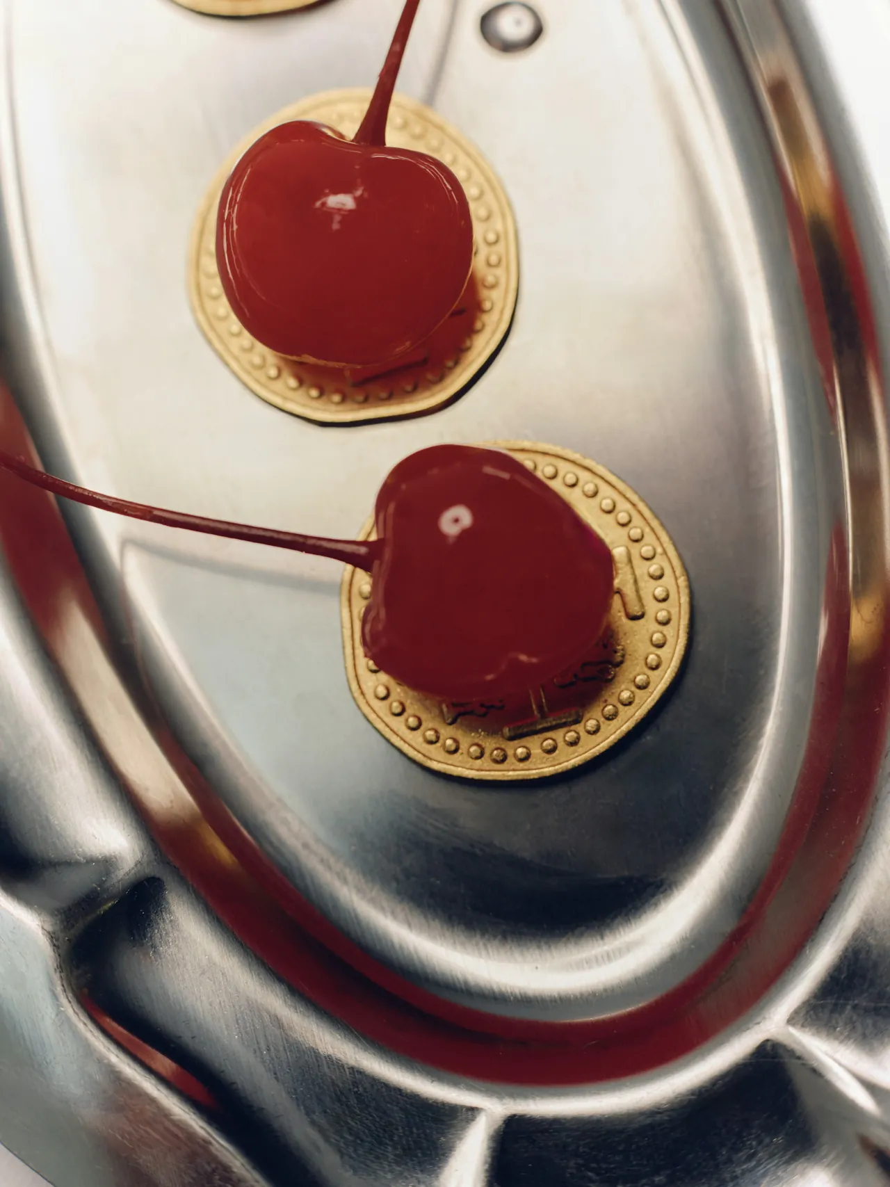 A close-up overhead shot of two glossy maraschino cherries with stems, each resting on a small embossed gold coin medallion inside a polished silver oval tray.