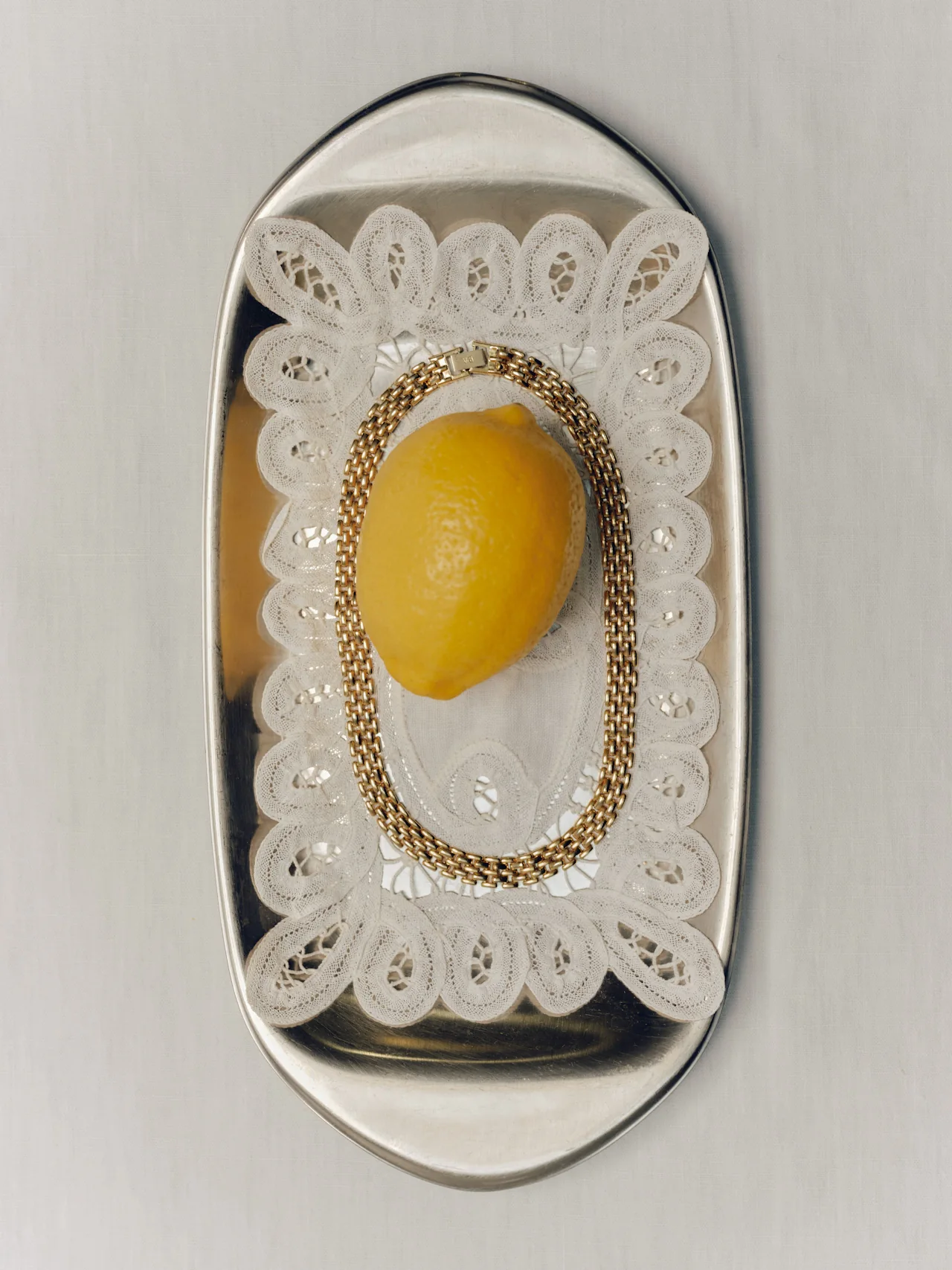 A polished silver oval tray holding a lemon wrapped in a gold chain bracelet, resting on a scallop-edged white lace doily, photographed from above against a grey surface.