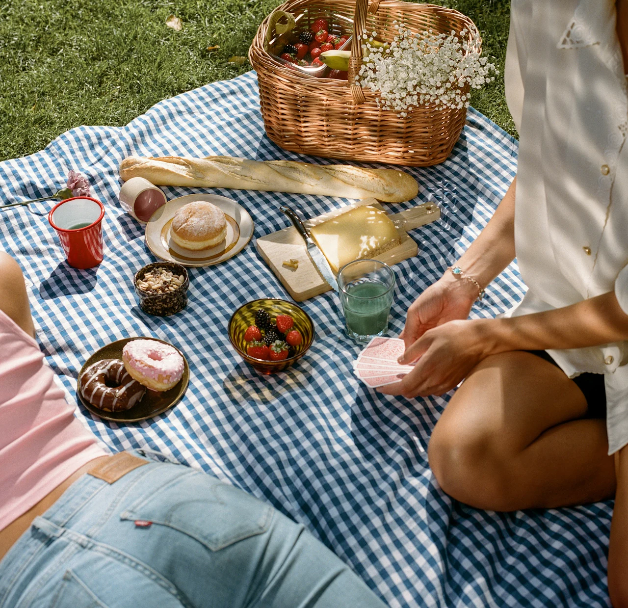 An overhead view of a sunny picnic spread on a blue gingham blanket, featuring donuts, baguette, cheese on a cutting board, fresh berries, granola, green drinks, and a wicker basket with baby's breath flowers. Two people are partially visible lounging and holding playing cards.