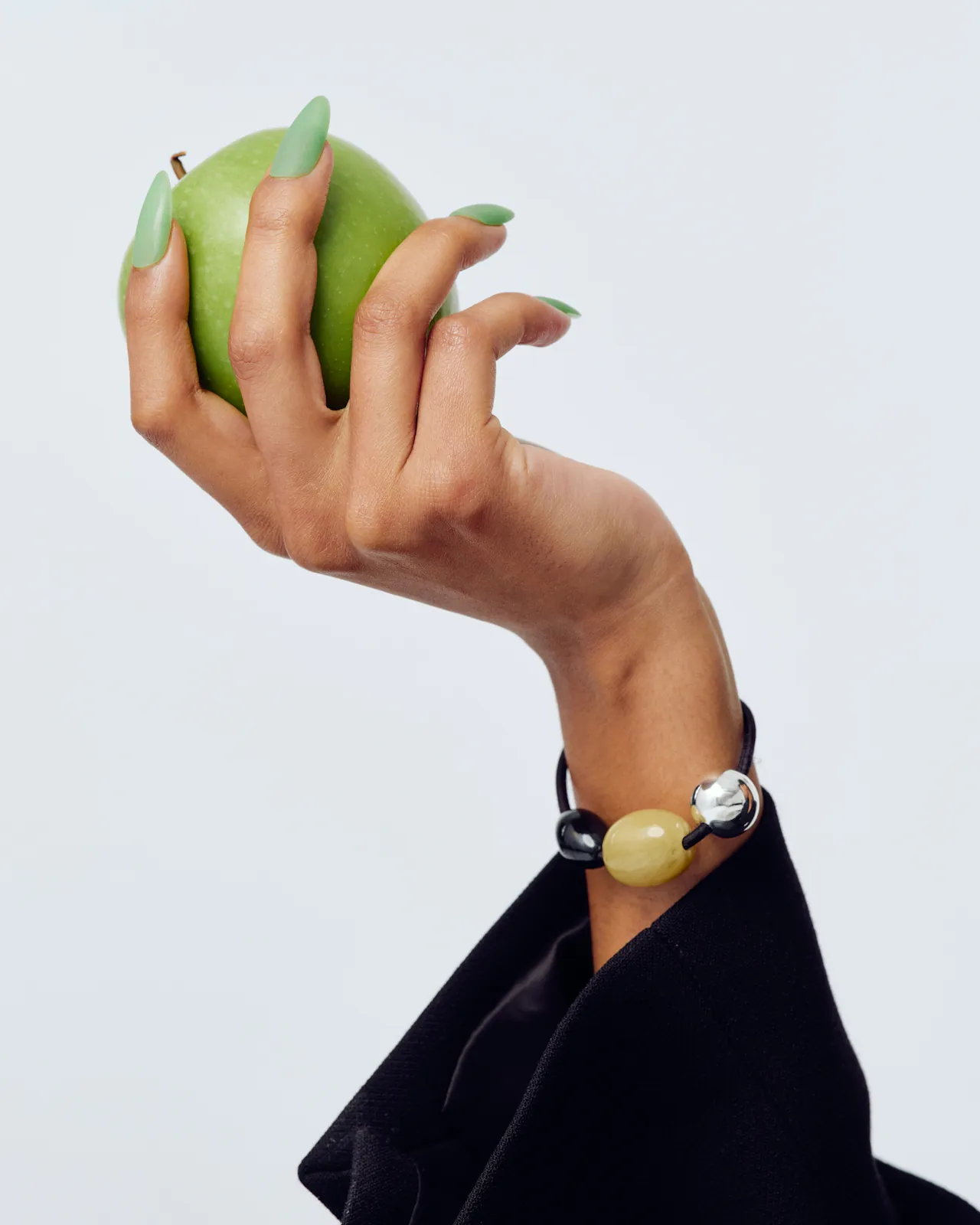A raised hand with long matte green nails grips a Granny Smith apple against a light grey background, wrist adorned with a chunky beaded bracelet in black, yellow, and silver, cuff of a black blazer visible.