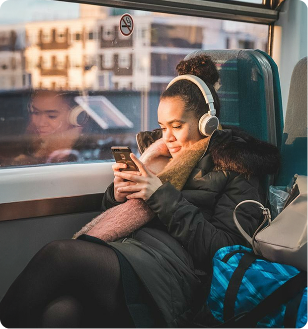 Woman wearing headphones and a scarf, sitting on a train seat while looking at her phone.