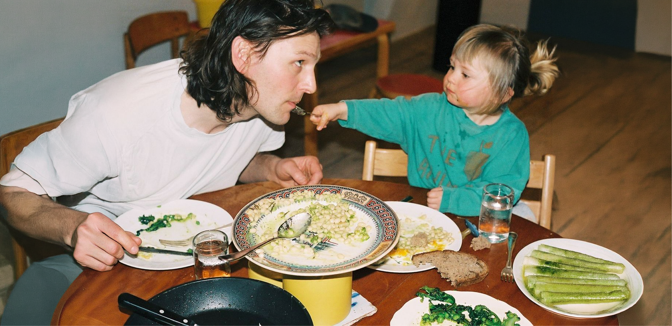 A young child in a teal shirt feeds a man with long hair sitting at a table with plates of food and a glass of water.