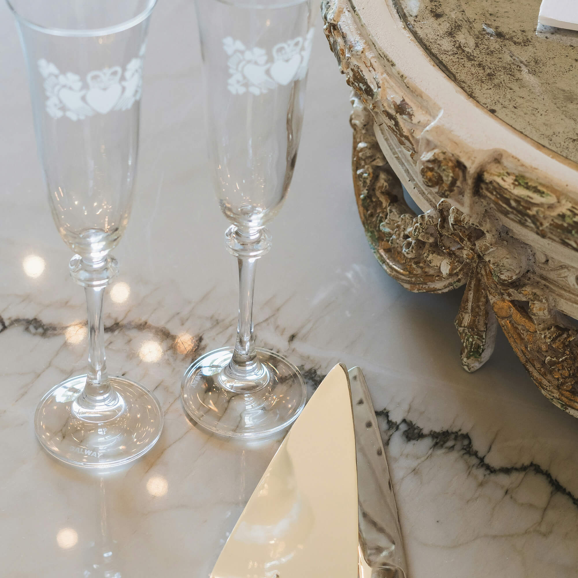 Two ornate champagne flutes next to a cake server on a marble surface beside a decorative cake stand.