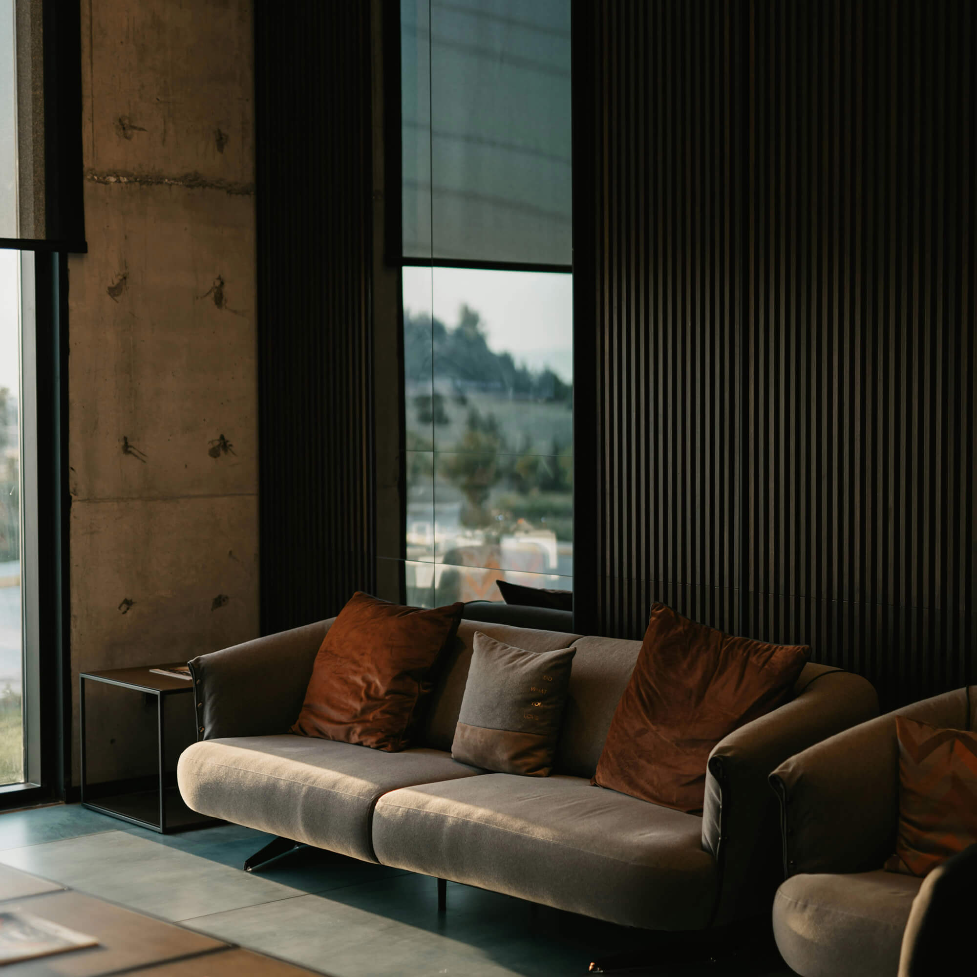 Modern living room with a grey sofa, brown cushions, a side table, and large windows letting in natural light.