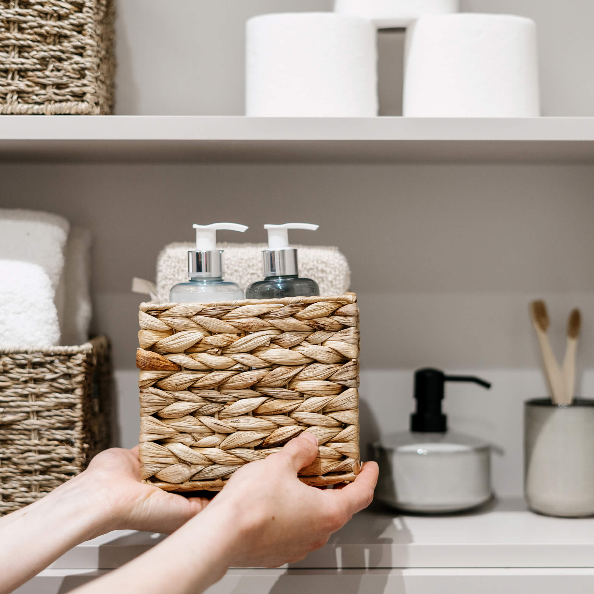 Person holding a woven basket with two pump bottles and a towel on a bathroom shelf.