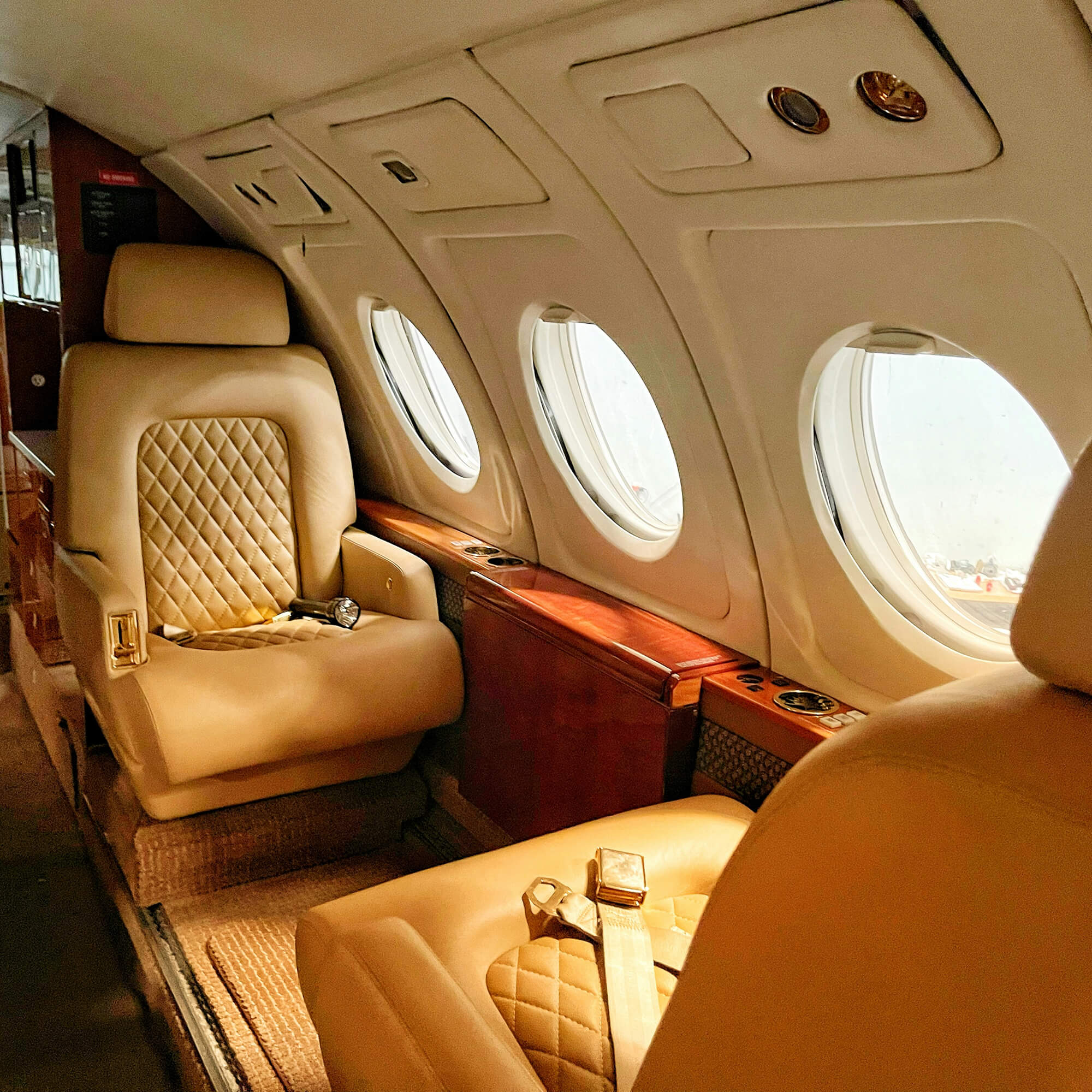 Interior of a private jet with beige leather seats, three oval windows, and wood paneling.