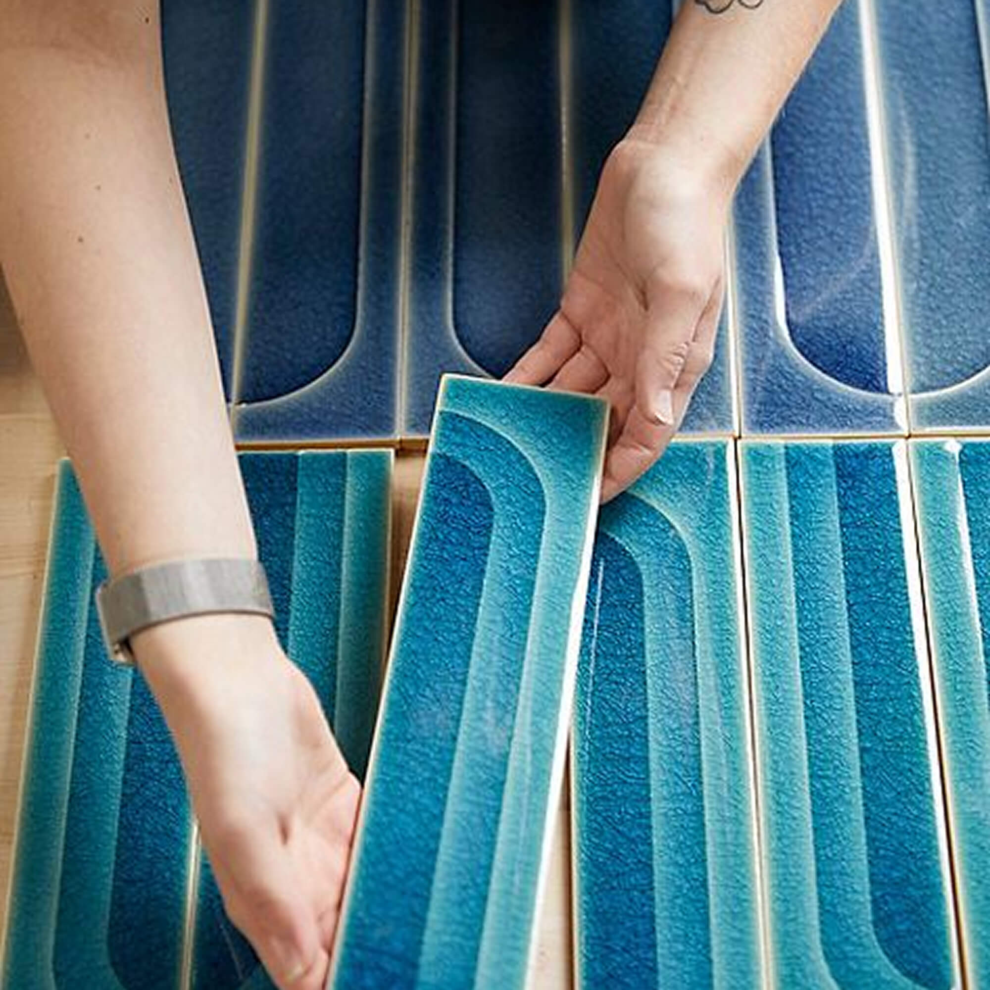 Hands arranging rectangular blue ceramic tiles with curved patterns on a wooden surface.