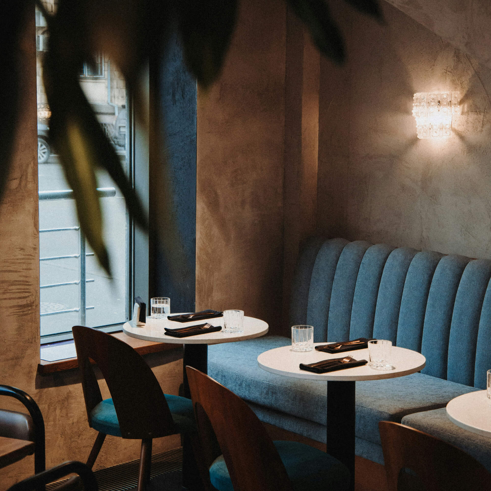 Cozy restaurant corner with round white tables, blue cushioned booth seating, and wooden chairs set with glasses and black napkins with silverware.