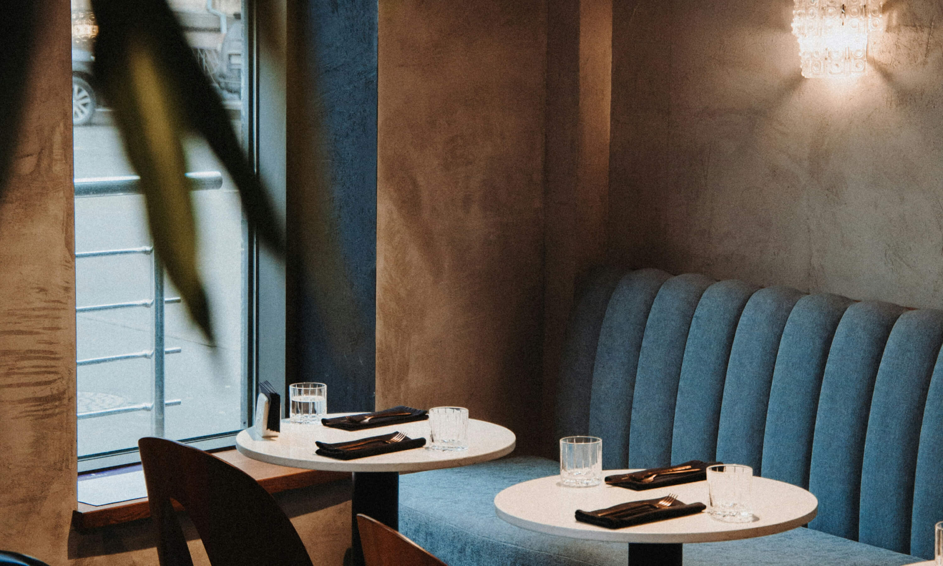 Cozy restaurant corner with two round white tables set with black napkins, forks, and glasses, next to a blue cushioned bench and a window.