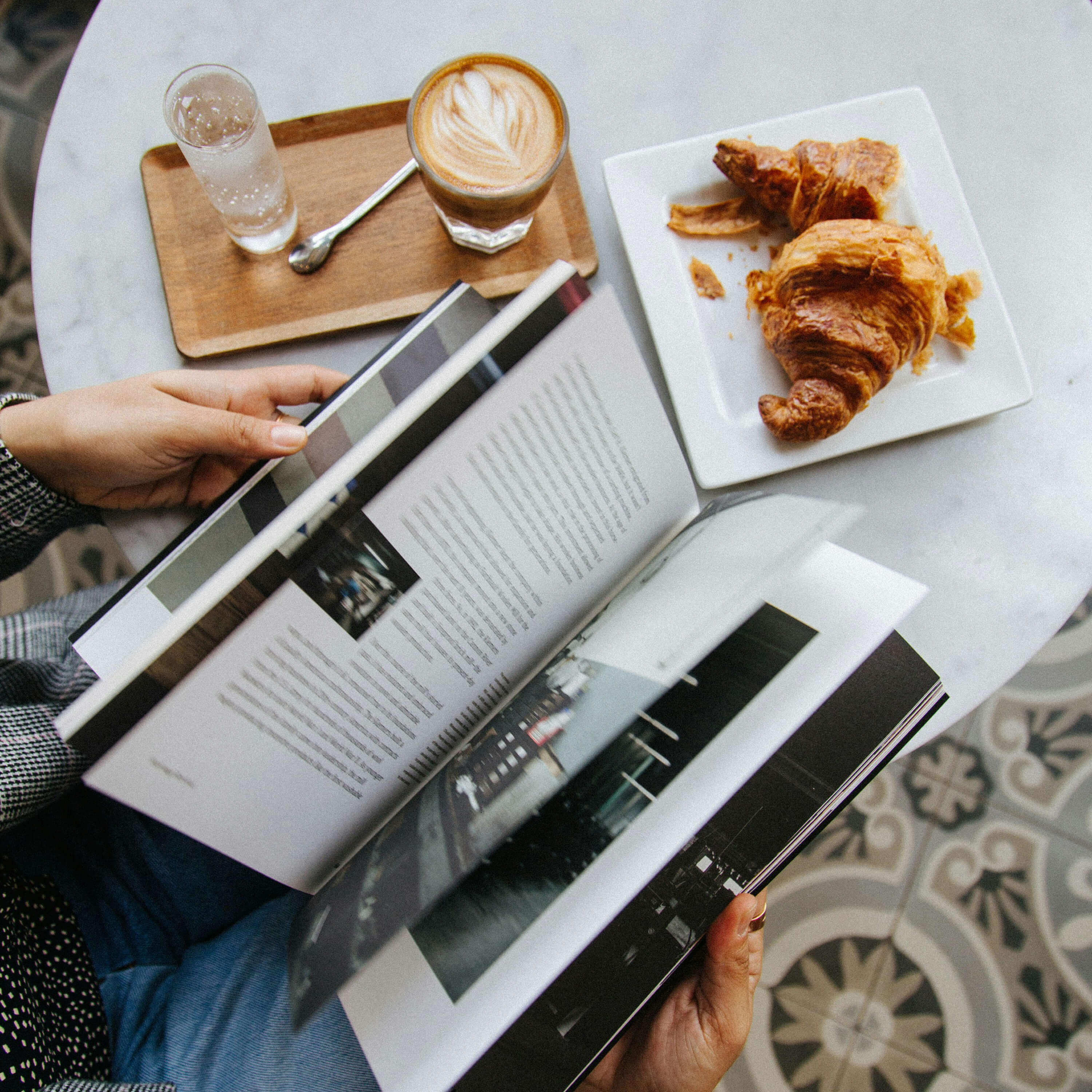Person reading a magazine at a marble table with a latte, water glass on a wooden tray, and a croissant on a white plate.