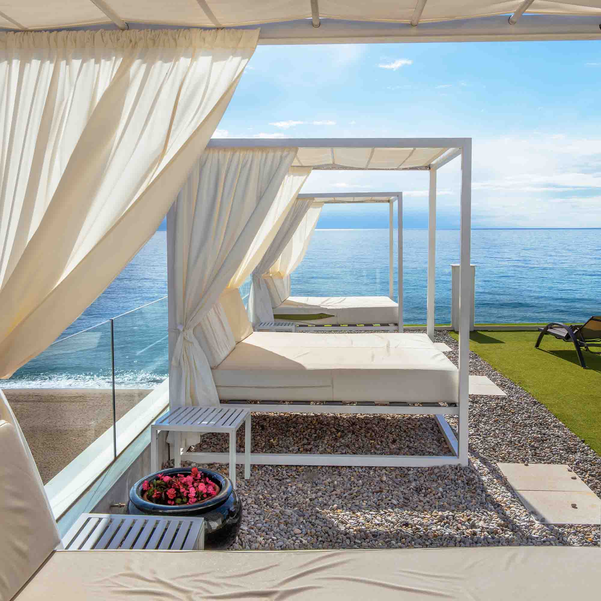 White canopy beds with curtains overlooking a calm sea under a bright sky.