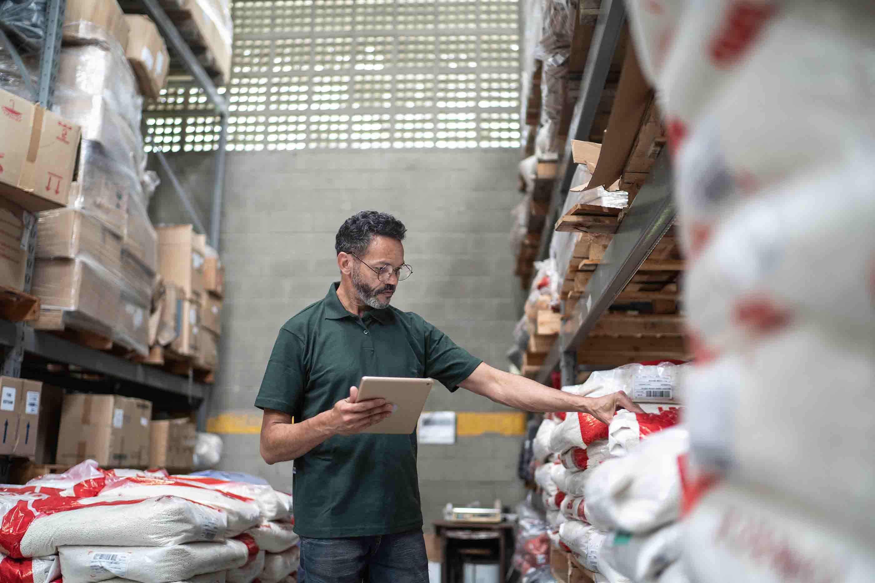 Man in a warehouse using a tablet to check inventory on shelves stacked with bags and boxes.