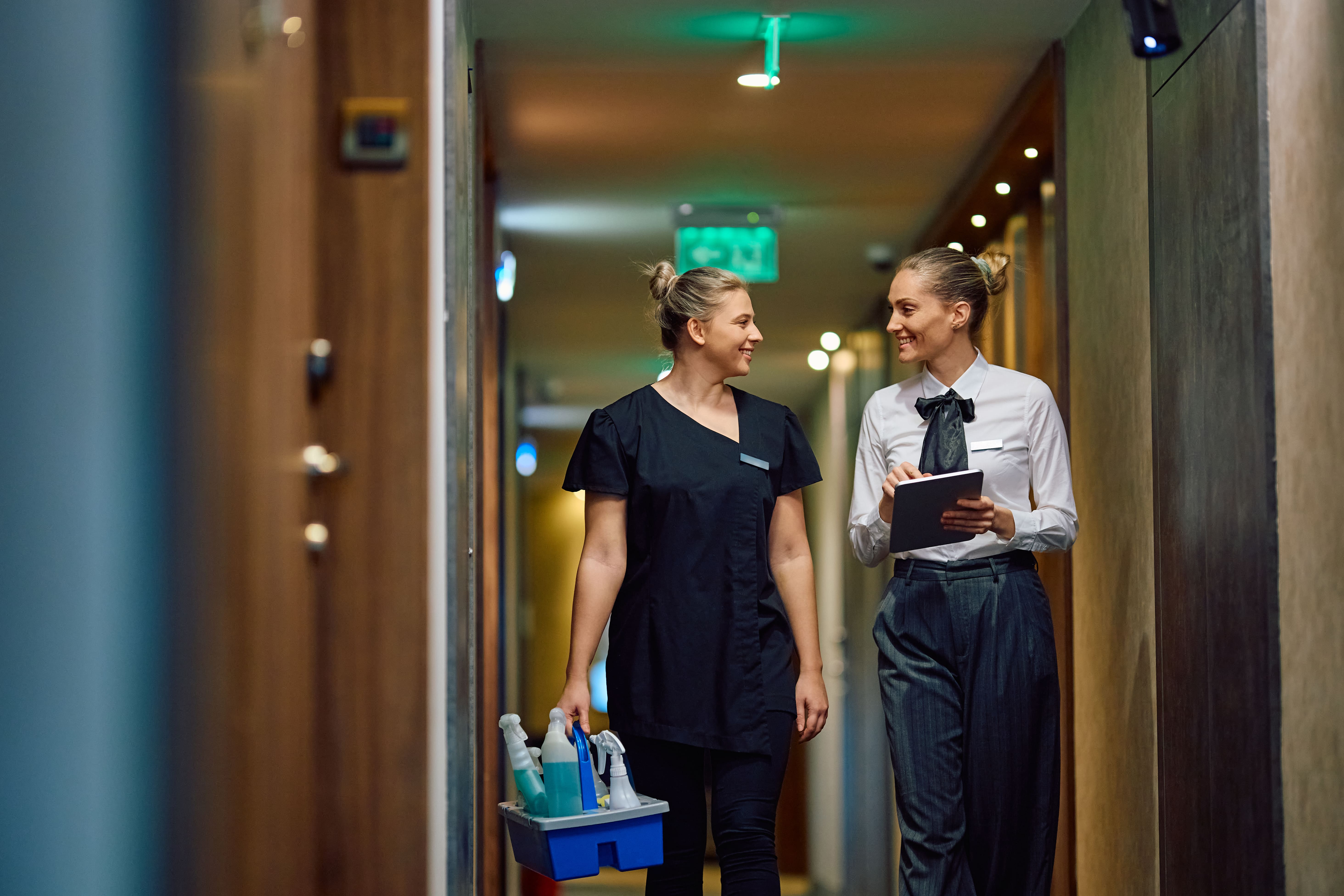 Two female hospitality staff walking in a hotel hallway, one holding cleaning supplies and the other using a tablet.
