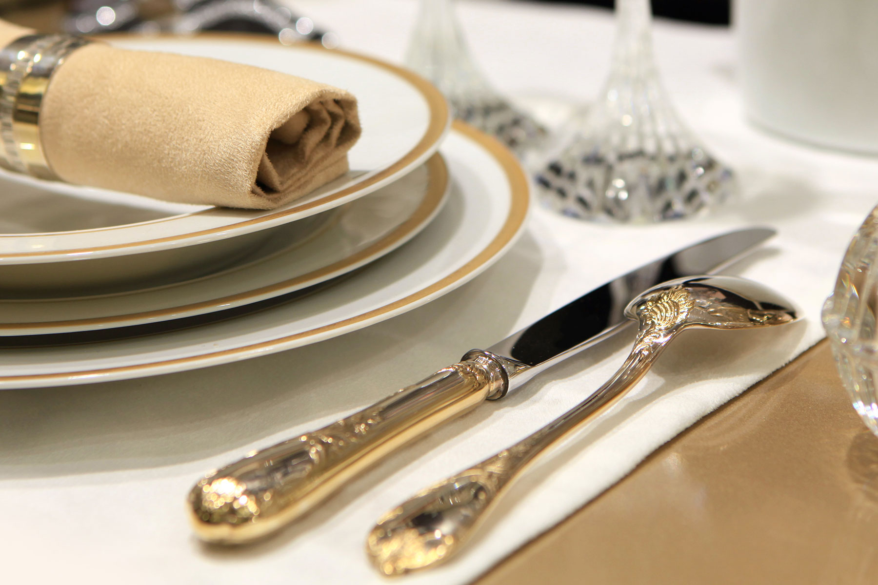Close-up of elegant table setting with stacked white plates with gold rims, beige rolled napkin, and ornate gold-handled knife and spoon on white tablecloth.