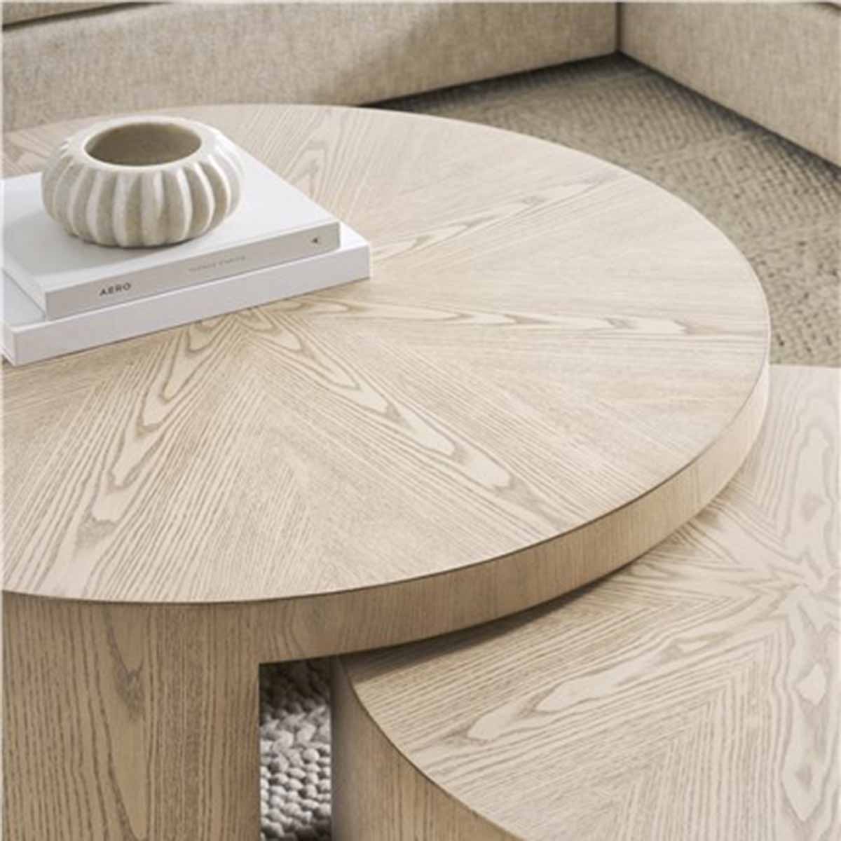 Close-up of two nested round wooden coffee tables with a light finish, one table topped with two stacked books and a decorative ceramic piece.