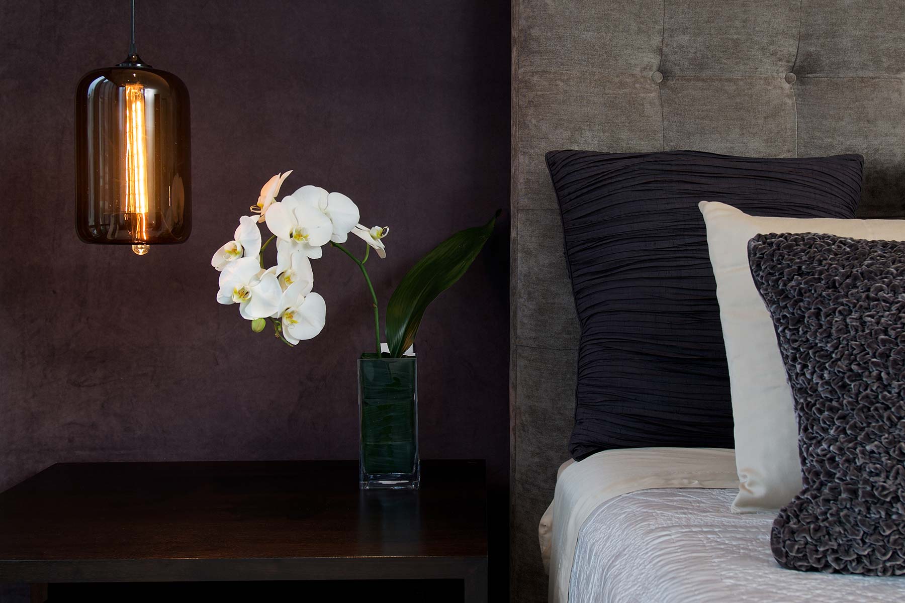 White orchid in a square glass vase on a dark wooden nightstand next to a bed with textured dark and light pillows.