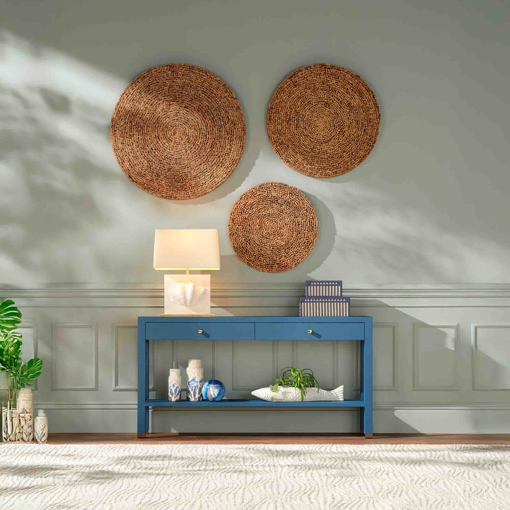 Blue console table with two drawers against a gray wall, decorated with three round woven wall hangings, a white table lamp, ceramic vases, a patterned box, and a white fish-shaped planter with greenery.