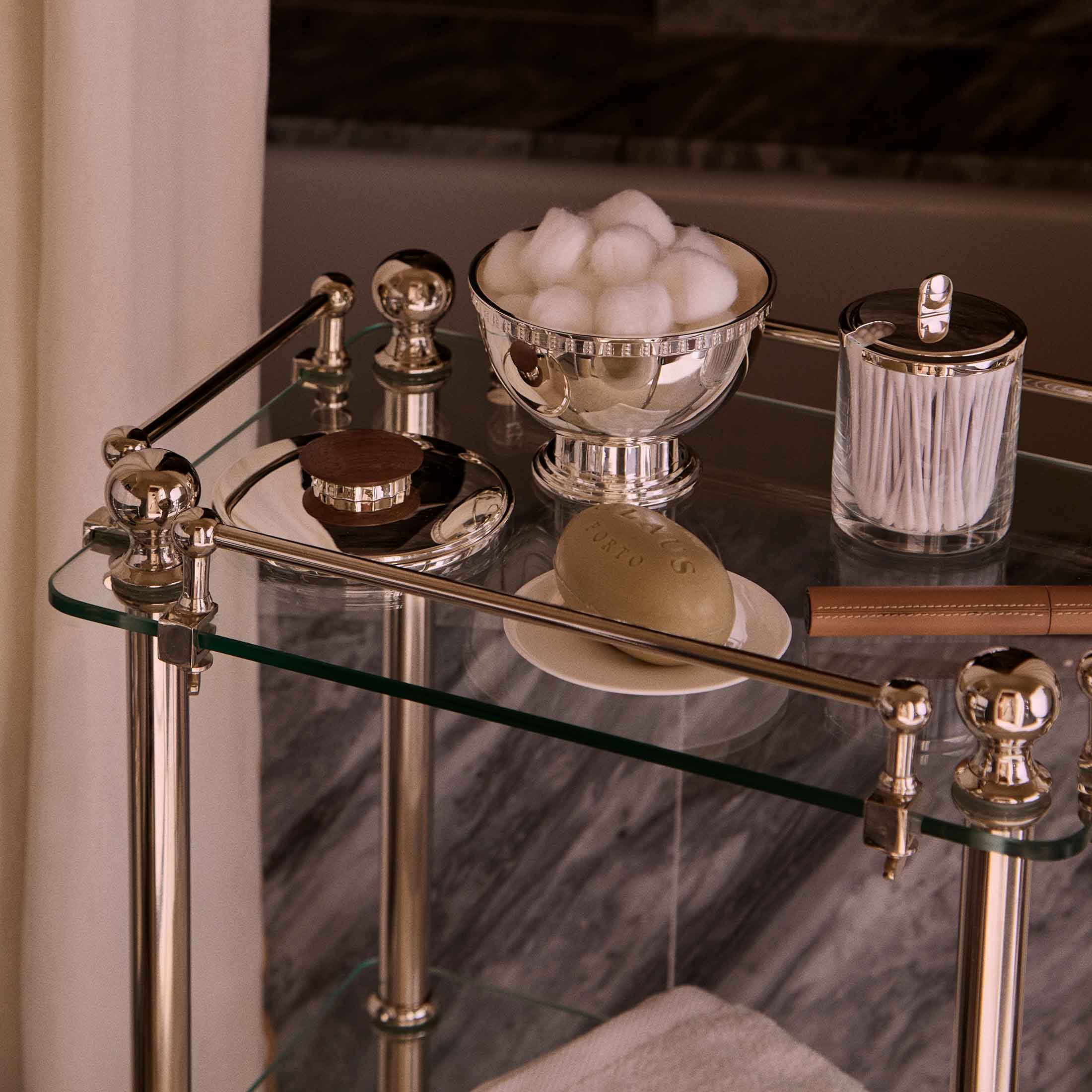 Puiforcat lass and chrome bathroom cart with cotton balls in a silver bowl, cotton swabs in a glass container, a bar of soap on a dish, and a leather-wrapped item.