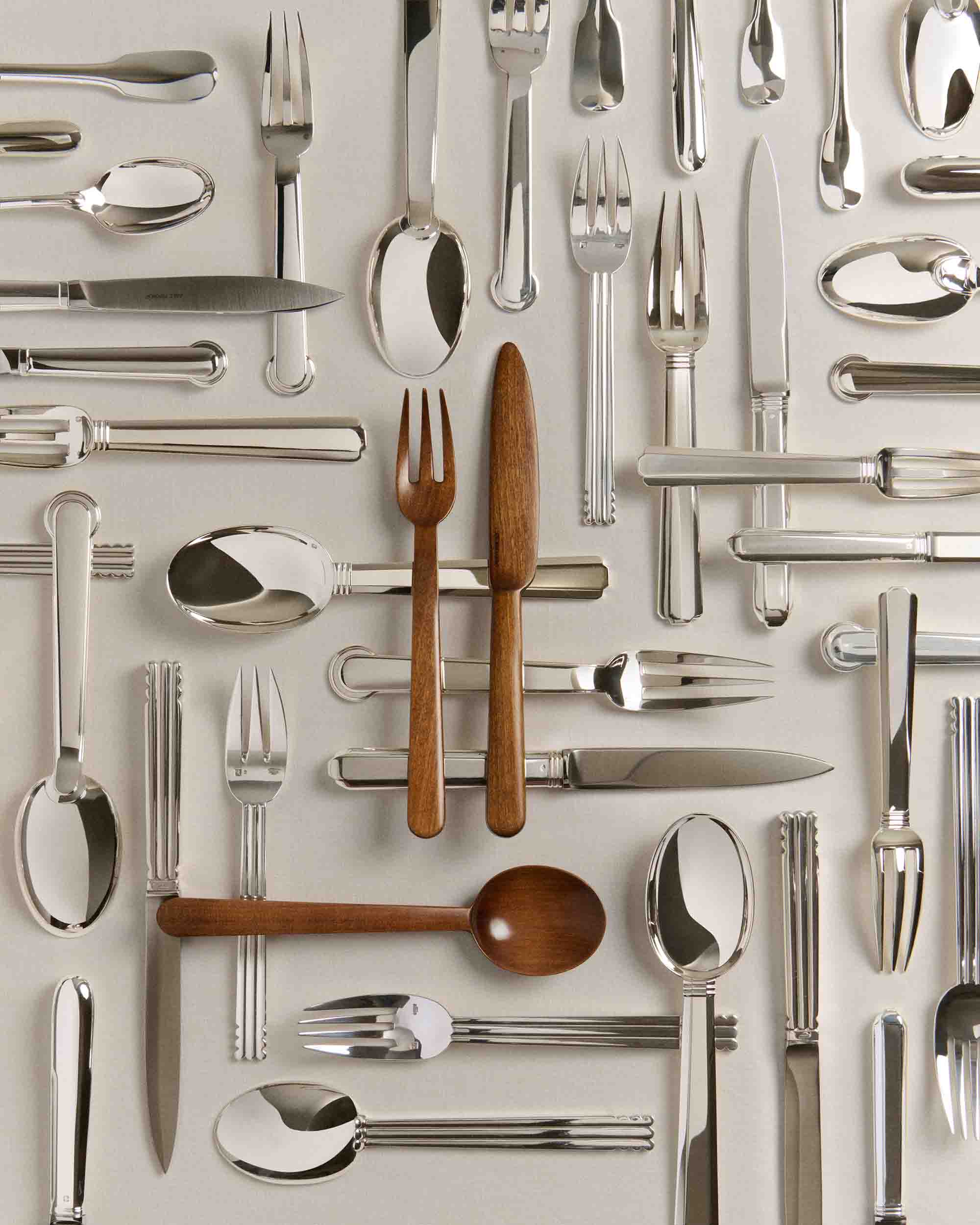 Assortment of Puiforcat silver metal forks, spoons, and knives with three wooden utensils arranged on a beige background.