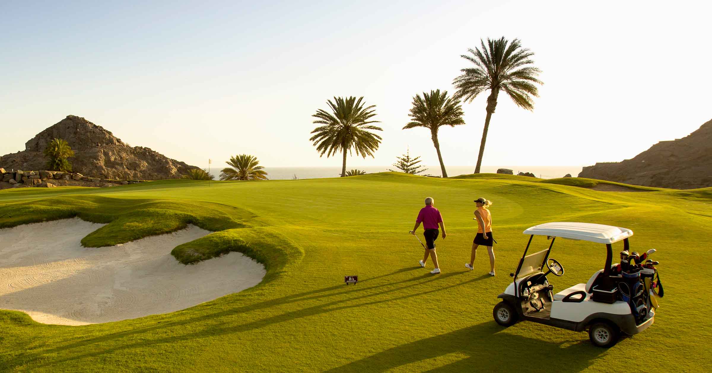 Two golfers walking on a green golf course near sand bunkers with palm trees and rocky hills in the background at sunset.
